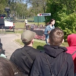Grupa uczniów słucha przewodnika, w tle stara lokomotywa wąskotorowa.