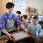 Grupa uczniów obserwuje proces czerpania masy papierowej z drewnianej balii.