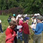 Duża grupa dzieci stoi na łące i rozmawia ze sobą. Chłopiec na pierwszym planie trzyma podkładkę z przypiętą kartką.