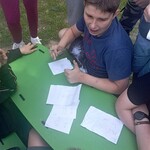 Chlopiec siedzi przy stoliku. Przed nim leżą kartki z rebusem, a wokół niego zgromadzeni są inni uczniowie.