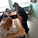 Pani policjantka stoi oparta o ławkę_ przy której dwoje uczniów rozwiązuje zadania znajdujące się na kartce.jpg