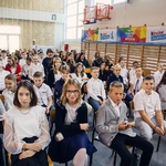 Duża grupa uczniów ubranych na galowo siedzi na krzesłach ustawionych na sali gimnastycznej.jpg