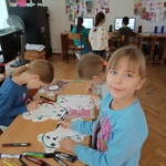 Trzy dziewczynki przy stoliku malują mazakami styropianowe szablony misiów. W tle kilkoro dzieci  pracuje przy komputerach.jpg