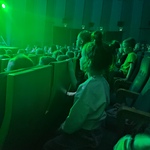 Dzieci siedzą w słabo oświetlonym audytorium i obserwują scenę oświetloną na zielono..jpg