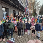 Grupa uśmiechniętych dzieci w zimowych kurtkach i czapkach stoi przed szarym budynkiem..jpg