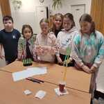 Grupa dzieci stoi przy stole_ prezentując swoją konstrukcję zbudowaną z papierowego kubka_ słomek i gumek recepturek..jpg