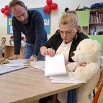 Miś Staszek siedzi przy stoliku z opiekunami świetlicy.jpg