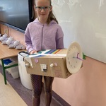 Dziewczynka stoi w klasie i trzyma własnoręcznie wykonany model wehikułu czasu_ ozdobiony zegarem i kolorowymi dekoracjami..jpg