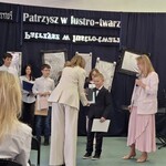 Pani dyrektor wręcza uczniowi dyplom. W tle inni uczestnicy konkursu na tle ściany udekorowanej imitacjami luster..jpg