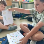 Chłopiec pokazuje rysunek z namiotem_ drzewem i słońcem_ obok ma ozdobione pudełko z pamiątkami..jpg