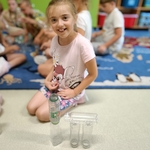 Dziewczynka w różowej koszulce uśmiecha się_ siedząc na podłodze i budując konstrukcję z plastikowych butelek..jpg