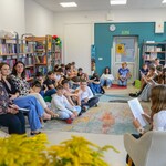 W bibliotece uczennica siedzi na fotelu i czyta. Naprzeciwko niej siedzą nauczyciele i  uczniowie..jpg