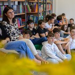 W bibliotece nauczyciele i uczniowie siedzą na krzesłach i dywanie. W tle półki z książkami. (2).jpg