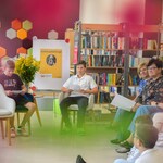 W bibliotece 2 uczniów siedzi na fotelach. Obok siedzą nauczyciele i uczniowie. W tle półki z książkami..jpg