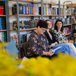 W bibliotece nauczyciele i uczniowie siedzą na krzesłach i dywanie. W tle półki z książkami..jpg