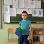 Chłopiec w turkusowej koszulce trzyma kolorową bryłę z klocków przed tablicą w klasie..jpg