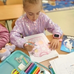 Dziewczynka w błyszczącym swetrze projektuje kropkową koszulkę. Obok leżą kredki i przybory szkolne..jpg