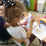 Dziewczynka w okularach i kokardzie w kropki wycina kolorowe kształty z kartki przy szkolnej ławce..jpg
