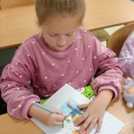Dziewczynka w różowej bluzie koloruje obrazek tajemniczy na kartce przy szkolnej ławce..jpg