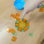 Palec wskazuje kolorowy wzór z klocków geometrycznych ułożony w okrąg na drewnianym blacie..jpg