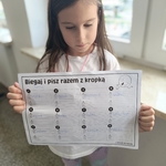 Dziewczynka w białej koszulce trzyma przed sobą kartę pracy Biegaj i pisz razem z kropką..jpg