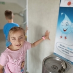 Dziewczynka w różowej koszulce stoi przy szkolnym poidełku obok plakatu z kroplą wody..jpg