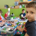 Chłopiec pokazuje zbudowaną konstrukcję z klocków_ za nim dzieci bawią się na dywanie..jpg