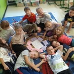 Grupa dzieci siedzi i leży na dywanie wokół rysunków przedstawiających plecak i różne przedmioty..jpg