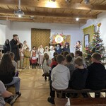 Uczniowie siedzący na ławeczkach oglądają tradycyjne przedstawienie jasełkowe z udziałem kolędników.jpeg