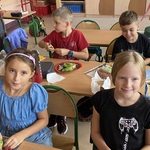 Na pierwszym planie przy ławce siedzą 2 uśmiechnięte dziewczynki_ za nimi dwójka chłopców robi kanapki.